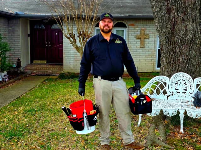 A smiling tech in uniform holding a tool bucket and a tool bag in a front yard