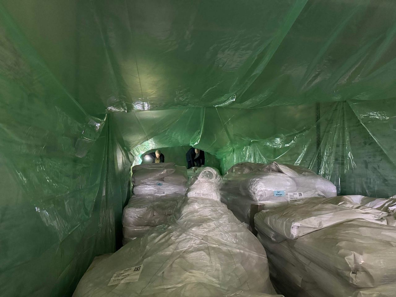 Rows of wrapped palletized bags inside a green tarp fumigation enclosure, two workers at the far end