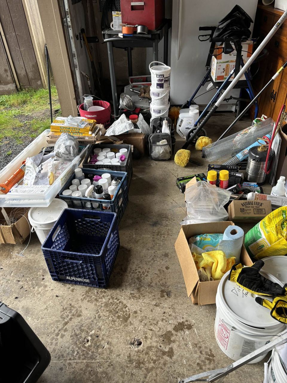 Garage floor crowded with milk crates of bottles, buckets, bags, and sprayer wands mid-reorganization